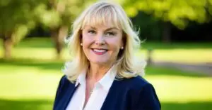 Smiling blonde woman in a navy blazer and white blouse posing outdoors in a sunny park.